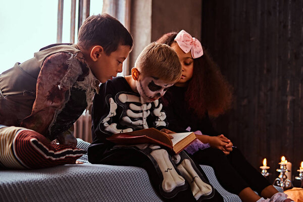 Три многорасовых ребенка в страшных костюмах читают страшные истории, сидя на кровати в старом доме
.