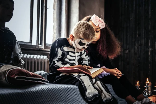 Tres niños multirraciales con disfraces aterradores leyendo historias de terror mientras están sentados en la cama en una casa vieja . —  Fotos de Stock