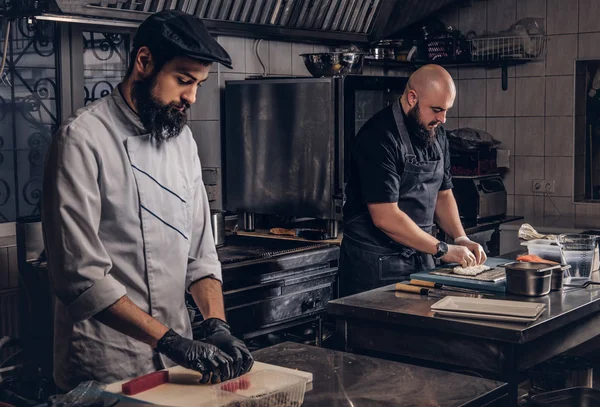 Dois cozinheiros brutais vestidos com uniformes preparando sushi em uma cozinha . — Fotografia de Stock