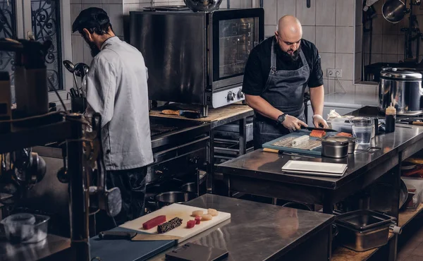 Dois cozinheiros brutais vestidos com uniformes preparando sushi em uma cozinha . — Fotografia de Stock