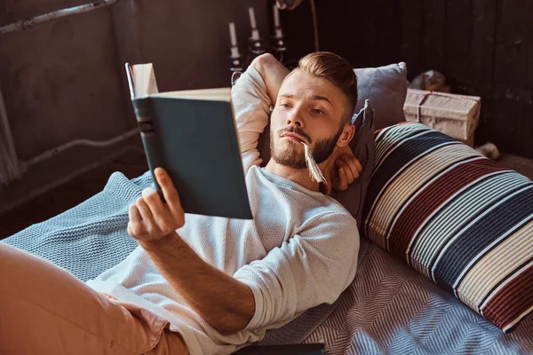 Portret przystojny student faceta czytanie książki i trzyma długopis w ustach, leżąc na łóżku. — Zdjęcie stockowe