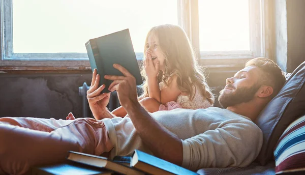 Όμορφος νεαρός πατέρας ανάγνωση παραμύθι η μικρή κόρη του στο κρεβάτι. — Φωτογραφία Αρχείου