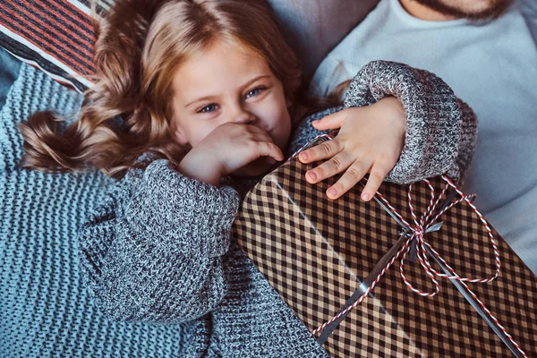 Retrato de cerca de una niña feliz en suéter caliente sostiene regalos mientras está acostada en la cama . — Foto de Stock