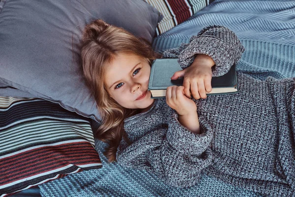 Close-up retrato de uma menina em camisola quente contém livro de histórias enquanto deitado na cama . — Fotografia de Stock