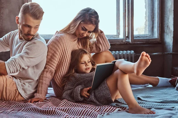 Mãe, pai e filha lendo livro de histórias juntos na cama . — Fotografia de Stock