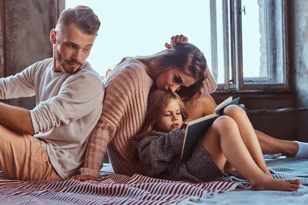 Mãe, pai e filha lendo livro de histórias juntos na cama . — Fotografia de Stock