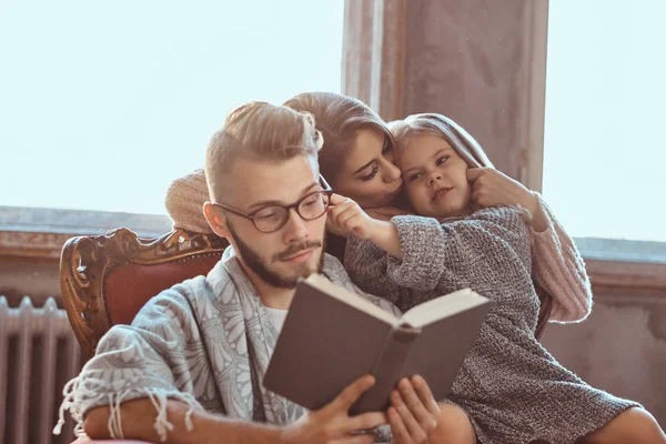 Reunião familiar. Mãe, pai e filha lendo livro de histórias juntos sentados no sofá. Conceito de família e paternidade . — Fotografia de Stock