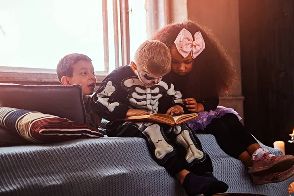 Tres niños multirraciales con disfraces aterradores leyendo historias de terror mientras están sentados en la cama en una casa vieja . —  Fotos de Stock