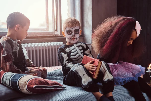 Три мультирасові діти в страшних костюмах читають історії жахів, сидячи на ліжку в старому будинку . — стокове фото