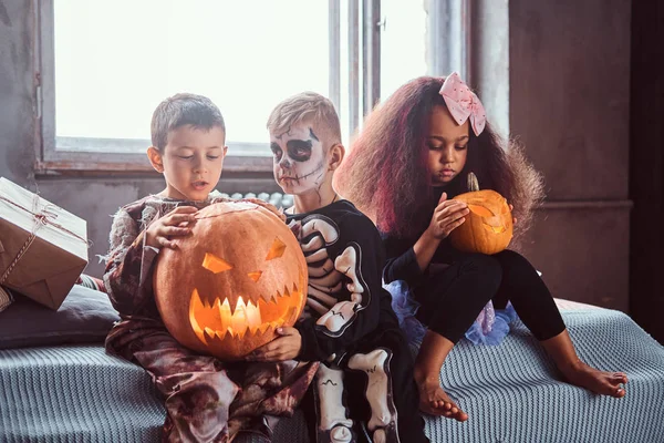 Grupo de niños multirraciales durante la fiesta de Halloween sostiene calabazas mientras está sentado en la cama en una casa vieja . — Foto de Stock