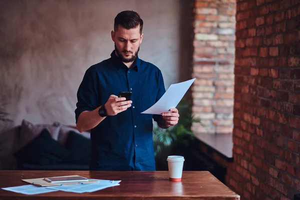 Hombre barbudo guapo usando un teléfono inteligente y trabajando con documentos de papel en la oficina con interior loft . —  Fotos de Stock