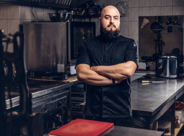 Mutfakta çapraz kolları olan siyah üniforma ayakta acımasız sakallı Şef aşçı.