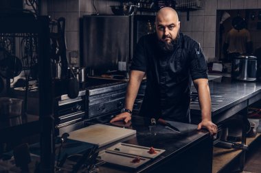 Bir masanın üzerine eğilerek ve mutfak kamerada bakarak siyah üniformalı acımasız Şef.