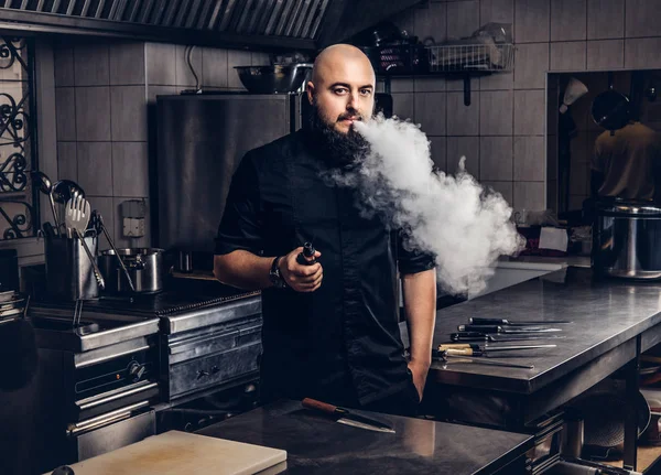 穿着黑色制服的大胡子厨师站在厨房里抽着电子烟. — 图库照片