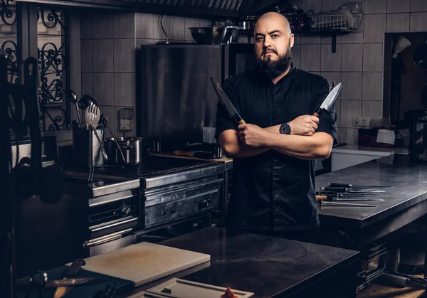 Cocinero barbudo en uniforme negro sostiene cuchillo y de pie con los brazos cruzados en la cocina . — Foto de Stock