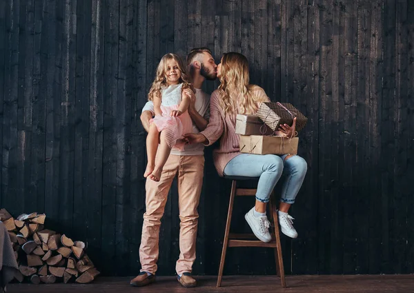 Concetto familiare. Bello padre tiene figlia sulle mani e baciare la moglie con doni in camera vuota contro un muro di legno . — Foto Stock