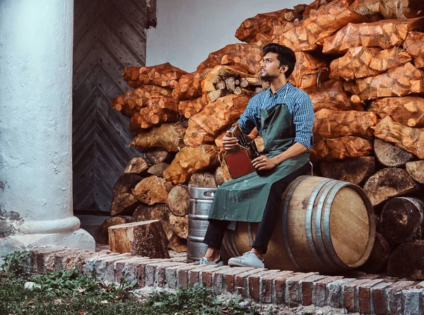 Cervecero cansado en delantal sentado en un barril y bebidas cerveza artesanal relajante después del trabajo . — Foto de Stock