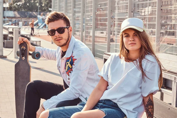 Trendi öltözött fiatal pár ül egy padon a városi sportkomplexum a napos, meleg tónusú gördeszka. — Stock Fotó