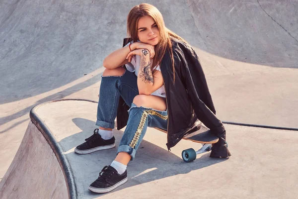 Güzel genç kaykaycı kız trendy bir longboard skatepark, oturan giymiş.