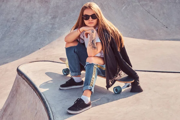 Joven chica hipster en gafas de sol vestida con jeans rasgados y camisa blanca sentada en un longboard en skatepark . —  Fotos de Stock
