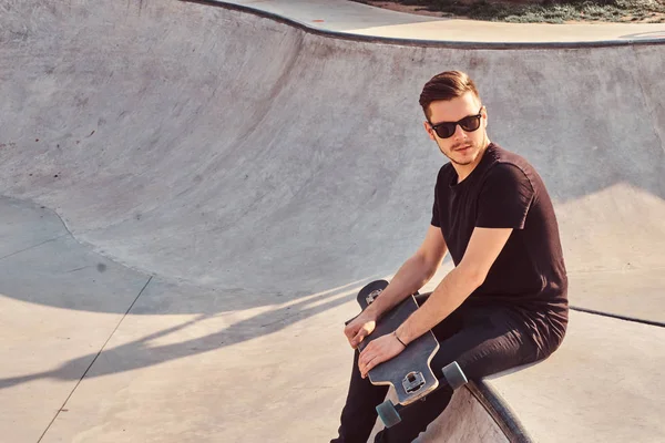 Stijlvolle hipster in zonnebril gekleed in zwarte jeans en shirt houdt skateboard zittend op skatepark. — Stockfoto