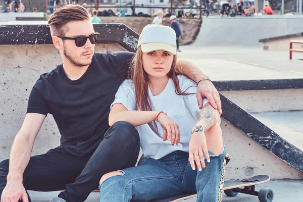 Trendi öltözött pár fiatal korcsolyázó átölelve együtt ülve a gördeszka a skatepark napsütéses napon. — Stock Fotó
