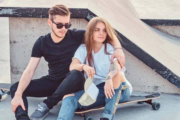 Trendy gekleed paar jonge schaatsers knuffelen elkaar zittend op skateboards in skatepark op zonnige dag. — Stockfoto