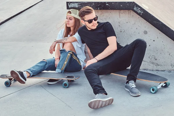 Pareja de patinadores jóvenes vestidos de moda sentados en monopatines en el skatepark . — Foto de Stock