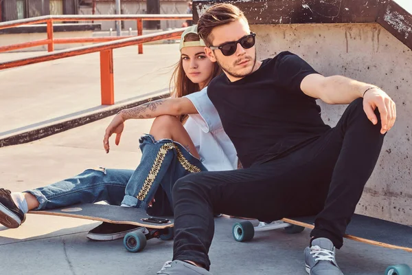 Trendiga klädd par unga åkare kramas tillsammans medan du sitter på skateboards på skatepark solig dag. — Stockfoto