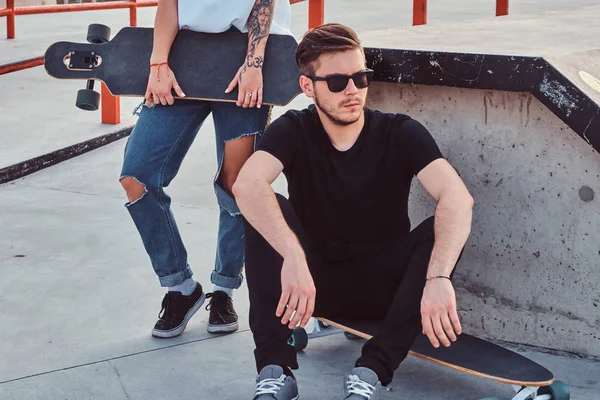 Porträtt av en snygg hipster kille sitter på en longboard på en skatepark. — Stockfoto