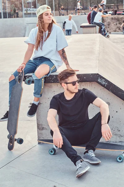 Joven pareja elegante de patinadores vestidos de moda que se relajan en un skatepark . — Foto de Stock