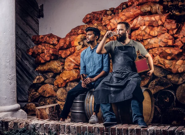 Brauer in Schürze und sein Freund sitzen zusammen auf einem Holzfass und trinken Craft Beer nach harter Arbeit in der alten Brauereifabrik. — Stockfoto