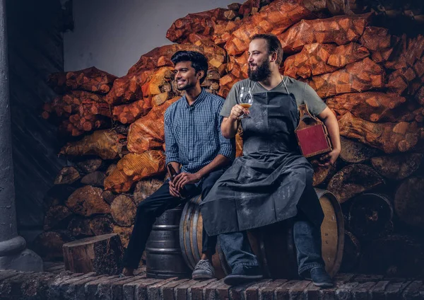 Brauer in Schürze und sein Freund sitzen zusammen auf einem Holzfass und trinken Craft Beer nach harter Arbeit in der alten Brauereifabrik. — Stockfoto