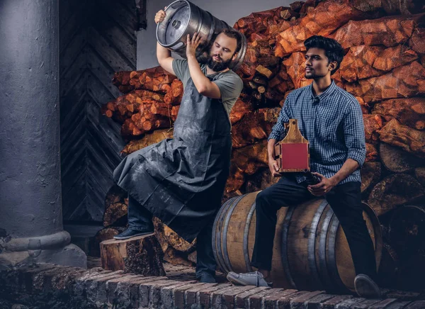 Cervecero barbudo en delantal sostiene barril y su amigo sentado en un barril de madera con cerveza artesanal en la fábrica de cerveza . — Foto de Stock