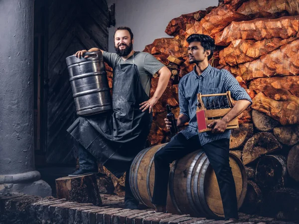 Cervecero barbudo en delantal sostiene barril y su amigo sentado en un barril de madera con cerveza artesanal en la fábrica de cerveza . — Foto de Stock