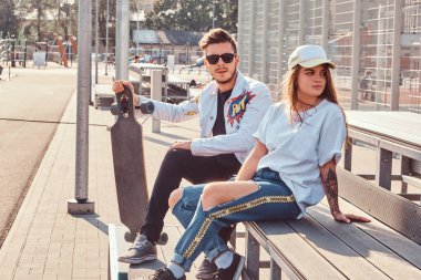 Atractive çift şık giyimli genç hipsters şehir spor kompleksi güneşli günde, bankta oturmuş kaykaylar ile.