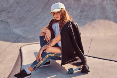 Yırtık kot pantolon ve beyaz gömlek bir longboard skatepark, oturan genç hippi kız şapkalı giymiş.