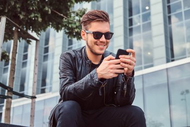 Gülümseyen güneş gözlüğü şık saç ile genç adam yakınındaki bir gökdelen otururken smartphone kullanarak siyah deri ceket giymiş..