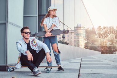 Şık giyimli çift - bir longboard ve kız arkadaşı açık havada duran üzerinde oturan genç hipster adam.