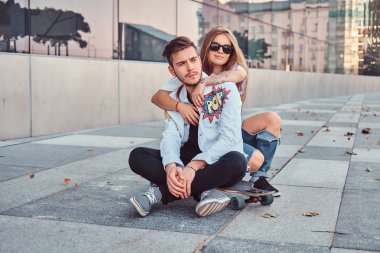 Genç çift trendy giyinmiş - güzel kız kucaklamak onlar birlikte gökdelen yakınındaki bir kaykay üzerinde oturan erkek arkadaşı.