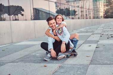 Genç çift trendy giyinmiş - güzel kız kucaklamak onlar birlikte gökdelen yakınındaki bir kaykay üzerinde oturan erkek arkadaşı.
