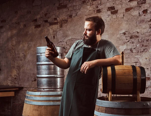 Cervecero en delantal de pie cerca de barriles y bebidas cerveza artesanal en la antigua fábrica de cerveza . — Foto de Stock