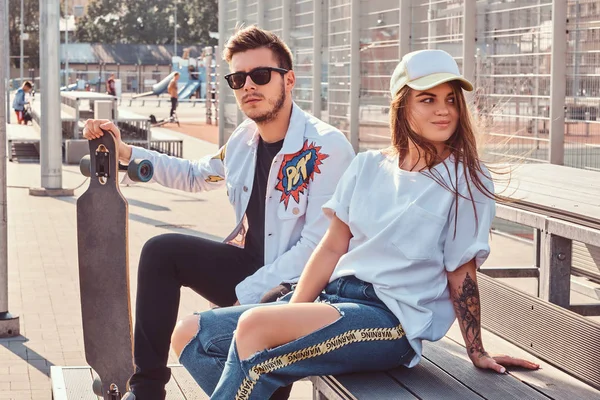 Atractive pár divatos öltözött fiatal hippik ül a padon a városi sportkomplexum, napsütéses napon gördeszka. — Stock Fotó