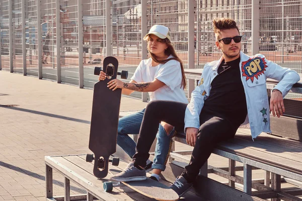 Trendy gekleed jong koppel met skateboards zittend op een bankje bij de stad sportcomplex op zonnige dag, met warm toned. — Stockfoto