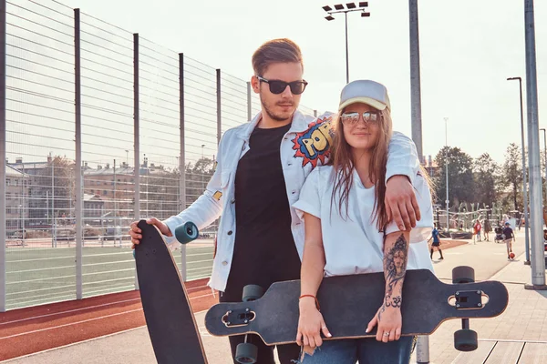 Tilltalande par trendiga klädda unga hipsters poserar med skateboards på city sport komplex solig dag — Stockfoto