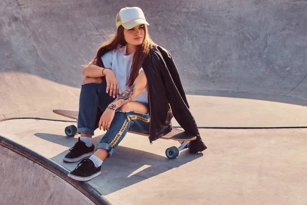Молодая хипстерша в кепке в разорванных джинсах и белой рубашке сидит на длинной доске в скейтпарке . — стоковое фото