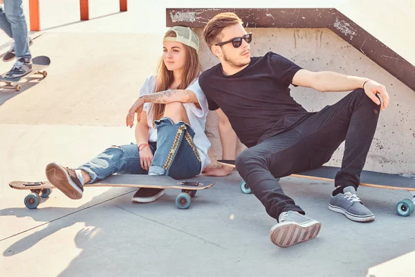 Trendi öltözött pár fiatal korcsolyázó átölelve együtt ülve a gördeszka a skatepark, napsütéses napon. — Stock Fotó