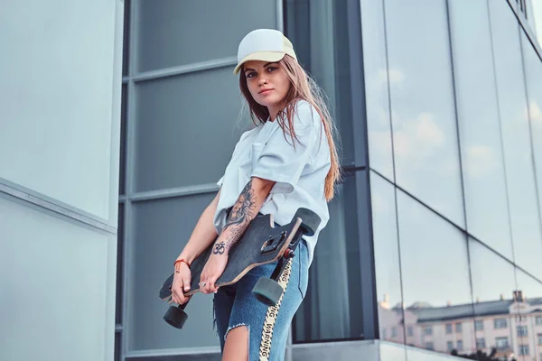 Giovane ragazza hipster in berretto vestito con una camicia bianca e jeans strappati tiene skateboard mentre posa vicino al grattacielo . — Foto Stock