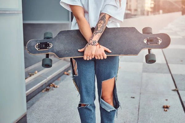 Cropped photo of a young hipster girl dressed in white shirt and ripped jeans holds skateboard while posing near skyscraper. — Stock Photo, Image