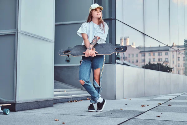 Menina jovem hipster em boné vestido com uma camisa branca e jeans rasgado mantém skate enquanto posando perto arranha-céu . — Fotografia de Stock